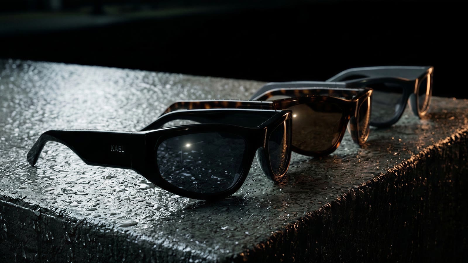 Three identical wraparound sunglasses in black, tortoiseshell and graphite on a wet concrete ledge